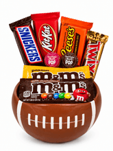 A football-shaped container filled with assorted branded chocolates and candy bars, including Snickers, KitKat, Reese's, Twix, M&M's, and Tootsie Roll Pop, on a white background.